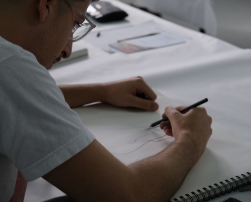 A person in glasses and white t shirt leaning on a notebook using their pencil to draw