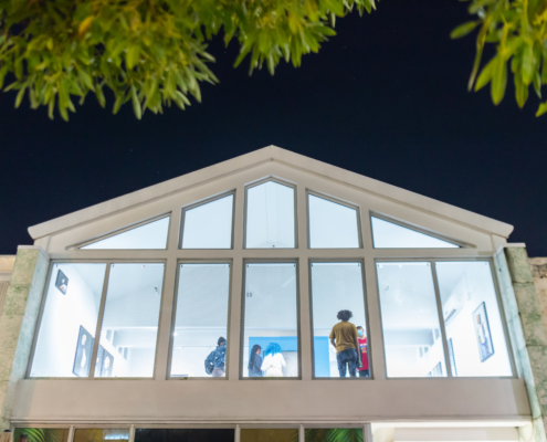 A white building with large glass windows show the illuminated interior of a gallery space at night. Green leaves from a nearby tree frame the photo.