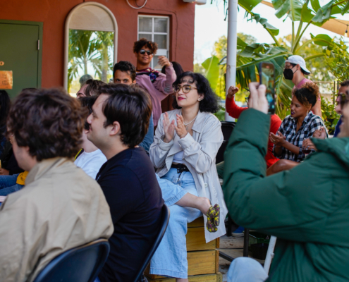 A group of people are seen sitting down and clapping during a gathering
