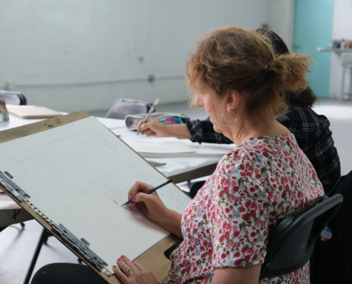 A person in a pink patterned top with reddish brown hair holds a pencil on a large clipboard in front of them.