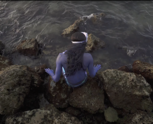 The back view of a violet colored merperson seated on the rocks