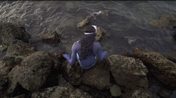 The back view of a violet colored merperson seated on the rocks