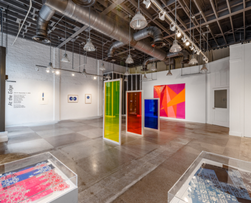 Wide view of a gallery with three neon colored sculptures in the center of the room. Two cases with artwork occupy the corner of the room