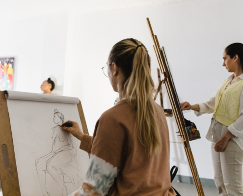 Long hair blonde person in a brown shirt facing an easel with pencil in hand
