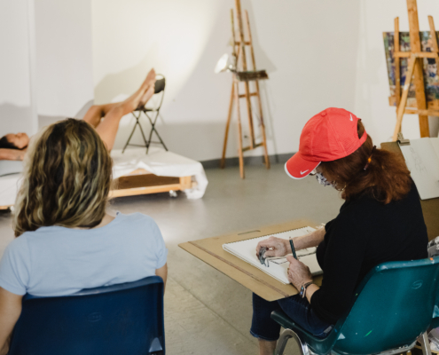 The back of the head of a person in blue seated next to person in red baseball cap drawing a person in front of them