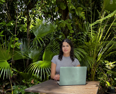 Amanda Bradley sits with a computer in front of tropical palm fronds