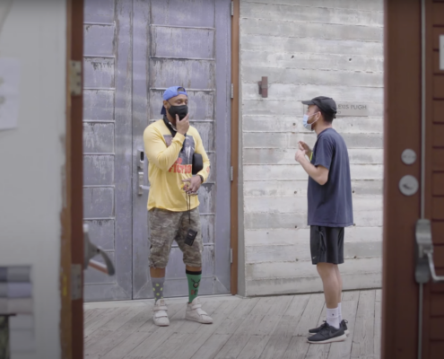Two artists stand in a outdoor area mid conversation. One is wearing a colorful outfit the other in neutrals