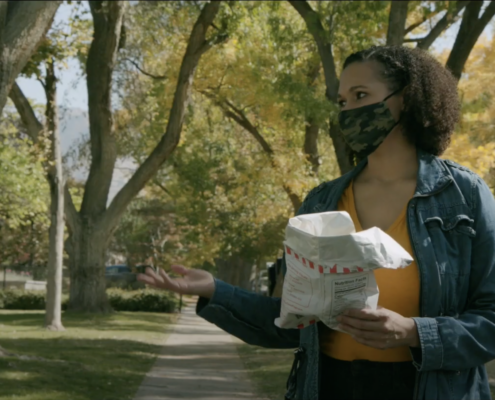 A person with neck length curly hair wearing a camo mask holding a bag of chips on the sidewalk surrounded by trees