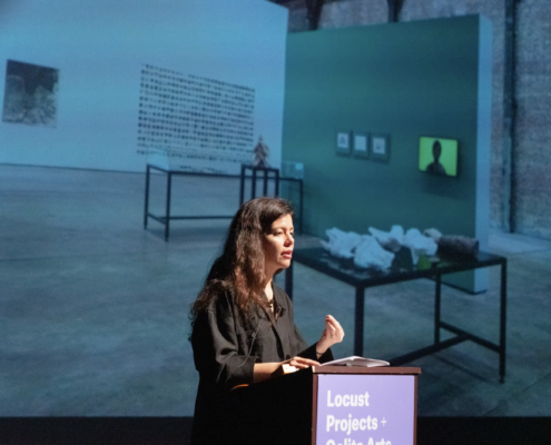 Ruba Katrib gesturing behind a podium with a photograph projected behind her