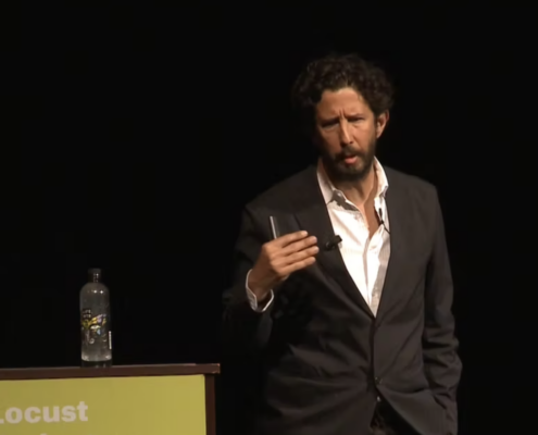 Jose Roca in a black suit with a unbuttoned white shirt standing next to a a podium with a water bottle at the top