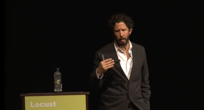 Jose Roca in a black suit with a unbuttoned white shirt standing next to a a podium with a water bottle at the top