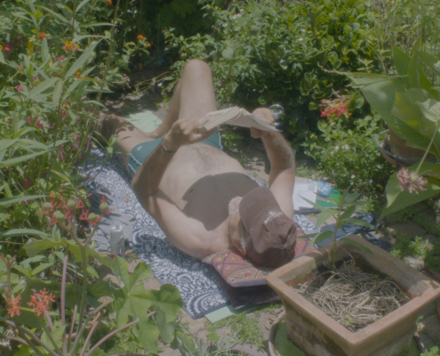 A shirtless person wearing a brown hat and green shorts lays on a patterned rug in the middle of a garden reading a book held over their head