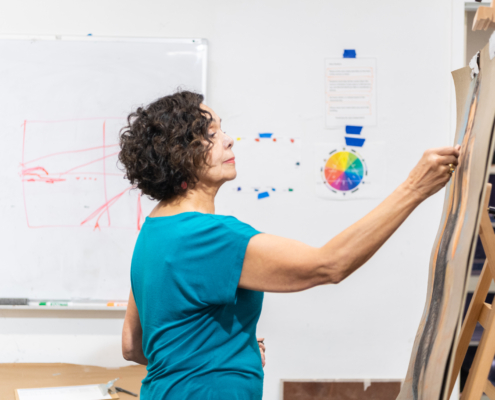 Brunette woman with short hair and a blue top stands profile in from of a butcher paper drawing with a white board behind filled with drawing instructions