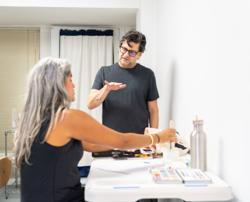 Raul Perdomo instruction a gray haired woman sitting at a desk with art supplies in use