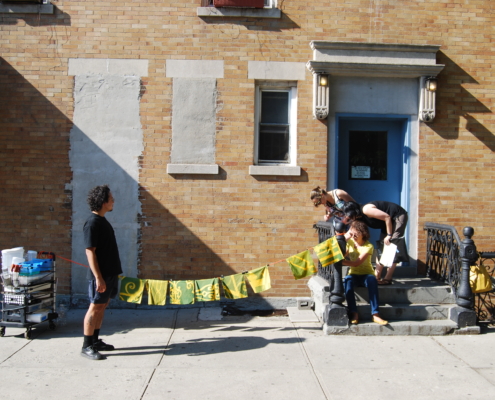 A person stands side profile next to a string of yellow square fabric pieces with various designs. the line is tied to a stoop with a few people engaging and looking at it.