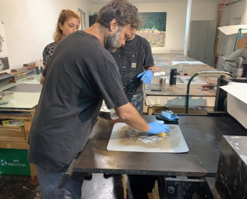 Person wearing blue gloves stands over a table with litho plate with two people partially obscured watch over.